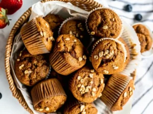 Overhead shot (4:3) of a rustic woven basket filled with numerous golden brown Simply Applesauce Muffins in white and natural brown paper liners, each topped with a scattering of rolled oats. The basket sits on a marble countertop, with glimpses of a white and grey striped cloth, a few fresh strawberries, and blueberries softly blurred in the background, under warm, natural morning light casting soft shadows. The scene is clean and tidy.
