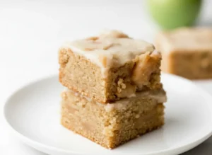 A stack of two Brown Butter Apple Blondies with creamy white glaze, showing the slightly crumbly texture and visible apple pieces. The blondies are a warm, light brown/caramel color. Shot on a minimalist white plate on a white marble countertop with soft natural morning light from an east window, soft shadows, warm tones, and a clean, tidy presentation. A subtle green blur is in the background. (4:3 ratio)