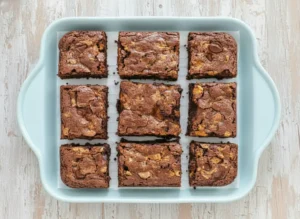 A top-down hero shot of freshly baked, fudgy Butterfinger brownies cut into perfect squares, showcasing their crinkly, dark chocolate top and visible lighter peanut butter candy swirls. The brownies are arranged on a minimalist white plate or a clean wooden cutting board, bathed in natural morning light from an east window, with soft shadows, warm tones, and a clean marble countertop in the background. (4:3 ratio)