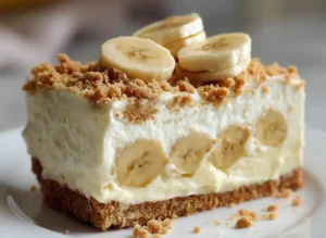 A generous slice of No-Bake Banana Pudding Cheesecake, clearly showing the distinct layers of graham cracker crust, fresh banana slices embedded in the creamy white filling, and a fluffy topping with more banana slices and graham cracker crumbs. The slice is on a minimalist white plate on a marble countertop with soft natural morning light from an east window, featuring warm tones and soft shadows. A wooden cutting board and fresh herbs are subtly blurred in the background, creating a clean and tidy presentation. No hands or people.