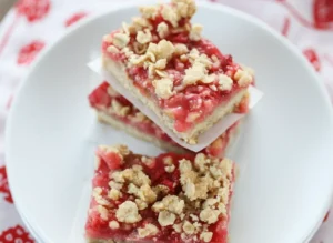 A stack of two square Strawberry Rhubarb Bars with a third bar beside them, all placed on a single light blue ceramic plate with white parchment paper underneath each bar. The bars feature a golden-brown oat crumble topping over a vibrant red strawberry rhubarb filling and a buttery shortbread crust. Shot with natural morning light from an east window, soft shadows, warm tones. Marble countertop with a subtle wood accent in the background. Clean and tidy presentation, no hands or people. (4:3 ratio)