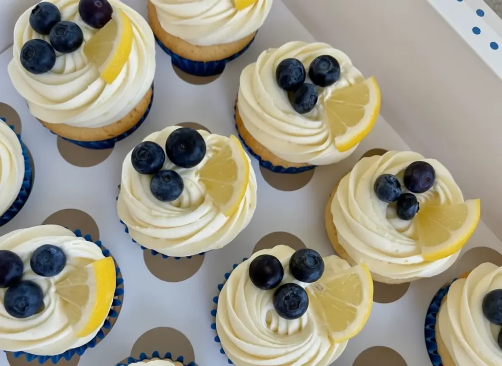A beautifully styled shot of one or two Blueberry Lemon Cupcakes on a minimalist white ceramic plate, featuring a swirl of white cream cheese frosting, topped with plump fresh blueberries and a vibrant lemon slice. The cupcakes are in fluted paper liners, sitting on a light marble countertop with a subtle wooden accent in the background. Natural morning light casts soft shadows, creating warm tones. (4:3 ratio)