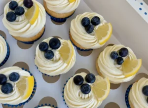 A beautifully styled shot of one or two Blueberry Lemon Cupcakes on a minimalist white ceramic plate, featuring a swirl of white cream cheese frosting, topped with plump fresh blueberries and a vibrant lemon slice. The cupcakes are in fluted paper liners, sitting on a light marble countertop with a subtle wooden accent in the background. Natural morning light casts soft shadows, creating warm tones. (4:3 ratio)