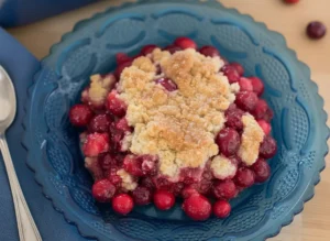 A beautifully baked cranberry cobbler in a white ceramic baking dish, with a single generous serving on a dark red decorative glass plate in the foreground. The cobbler features a golden-brown, crumbly topping and vibrant red, bubbling cranberry filling. Fresh cranberries and a sliced orange with a wooden reamer are artfully arranged in the soft-focused background. The scene is set on a light wooden surface with marble accents, illuminated by natural morning light, creating warm tones and soft shadows. The presentation is clean and tidy, reflecting a genuine love for the process, no hands or people.