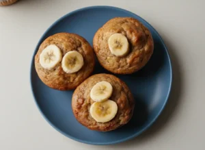 A beautifully plated close-up of several golden-brown Greek Yogurt Banana Muffins on a minimalist white ceramic plate. Each muffin is perfectly domed and topped with a few fresh, thin banana slices. The muffins have a tender, moist appearance and a slight golden sheen. The scene is bathed in natural morning light from an east window, casting soft shadows. A hint of marble countertop and a warm-toned wooden accent are visible in the soft-focus background, with the overall presentation being clean and tidy. No hands or people. (4:3 ratio)