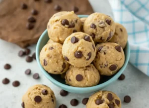 A high-quality 4:3 hero shot of several tan, dough-like protein bites studded with dark chocolate chips, stacked artfully in a small cream-colored ceramic bowl. The bowl is placed on a white marble countertop next to a rustic wooden cutting board. Soft, warm morning light streams in from an east window, creating gentle shadows. The background is clean and minimalist, featuring a small jar of fresh mint sprigs in a glass of water. The presentation is tidy and emphasizes the matte, doughy texture of the bites.