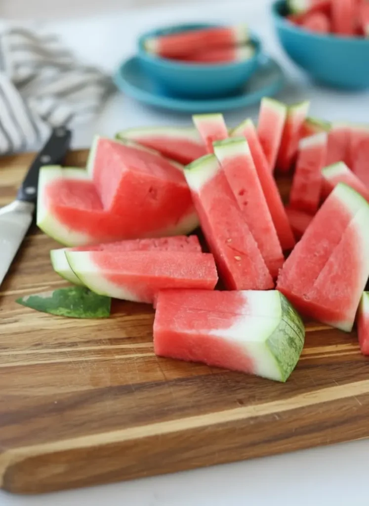 how to cut watermelon sticks - Ingredients and prep focus: A large, ripe watermelon sitting next to a sharp chef's knife on a classic wooden cutting board on a clean marble countertop, bathed in natural morning light. Fresh mint sprigs are artfully placed nearby, with soft shadows and warm tones. No hands or people.