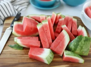 Hero shot of vibrant red watermelon sticks, some with a thin strip of green rind, artfully arranged on a minimalist white oval plate. Fresh green mint sprigs are scattered around the plate. The scene is set on a light marble countertop with warm natural morning light streaming from an east window, casting soft shadows. A corner of a striped kitchen towel is visible in the background, along with a subtle wooden accent. The presentation is clean and tidy.