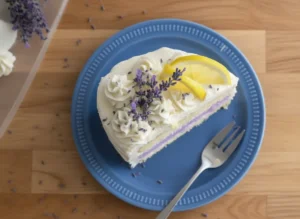 A hero shot of a three-layer cake on a minimalist white ceramic plate. The cake has two yellow layers and one vibrant purple layer in the center. It is covered in smooth white frosting with horizontal textured ridges on the sides. The top is decorated with white frosting rosettes, several fresh purple lavender sprigs, and two thin yellow lemon slices. The scene is set on a white marble countertop with soft morning light and a wooden cutting board partially visible in the background. 4:3 aspect ratio.