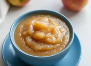 A minimalist white ceramic bowl filled with golden-brown, chunky stovetop applesauce, with visible cinnamon flecks and apple pieces. The bowl is on a simple white saucer on a light marble countertop. Blurred red apples are in the background. The scene is bathed in soft, natural morning light from an east window, casting gentle shadows. The presentation is clean and tidy, embodying a lived-in kitchen with genuine love for the process. (4:3 ratio)