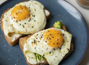 A high-angle shot of 10-Minute Avocado Egg Toast on a white ceramic plate. Two slices of dark, seeded multi-grain toast are topped with vibrant green smashed avocado and two sunny-side-up eggs with glistening, deep orange runny yolks. The eggs are generously coated in black and white sesame seeds, poppy seeds, and red pepper flakes. The plate sits on a white marble countertop with soft morning light streaming in from the side, creating gentle shadows. A small sprig of fresh cilantro is visible in the soft-focus background.