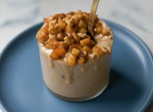 A hero shot of Apple Pie Overnight Oats in a clean, straight-walled glass on a white marble countertop. The oats are a creamy tan color, topped with a generous mound of sautéed, glistening cinnamon-coated apple chunks and chopped walnuts. A golden spoon is tucked into the side. Soft morning light from an east-facing window creates gentle shadows. In the background, a small wooden cutting board and a sprig of fresh mint in a ceramic vase are softly blurred. 4:3 aspect ratio.