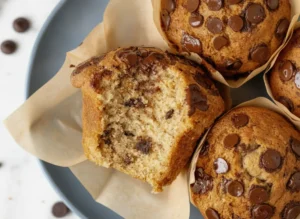A professional hero shot of three Bakery-Style Banana Chocolate Chip Muffins with high, golden-brown domed tops, heavily studded with melted semi-sweet chocolate chips, wrapped in brown tulip parchment liners, sitting on a white marble countertop with soft morning light and a wooden cutting board in the background, 4:3 aspect ratio.