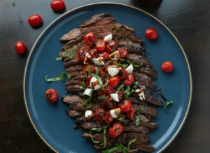 A hero shot of Balsamic Grilled Flank Steak Caprese sliced thinly on a minimalist white ceramic platter. The steak shows dark, caramelized grill marks and a pink medium-rare center. It is topped with bright red blistered cherry tomatoes, white mozzarella pearls, and vibrant green basil ribbons, all drizzled with a thick dark balsamic glaze. The platter sits on a white marble countertop with a wooden cutting board nearby. Soft morning light from an east window creates gentle shadows and warm tones. 4:3 aspect ratio.