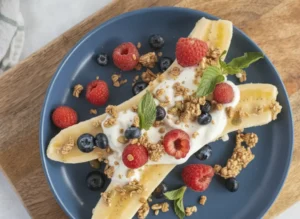 A high-angle hero shot of a Banana Split With Berries and Greek Yogurt on a minimalist white plate. The banana is split lengthwise with thick white Greek yogurt in the middle, topped with red raspberries, dark blue blueberries, and golden granola with pumpkin seeds. The plate sits on a rustic wooden cutting board over a white marble countertop. Soft, warm morning sunlight streams in from the side, creating gentle shadows. Fresh mint leaves are scattered as a garnish. A small ceramic bowl of granola is visible in the corner.