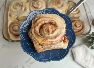 A high-angle 4:3 hero shot of a single, large Orange Cinnamon Roll on a white scalloped ceramic plate. The roll features prominent dark cinnamon swirls and is covered in a thick, glossy white glaze with visible bright orange zest specks. The plate sits on a white marble countertop with a soft wooden cutting board partially visible in the background. Natural morning light creates soft shadows and a warm, inviting atmosphere. Minimalist holiday decor like a small candle and a tiny green tree are blurred in the background.