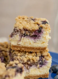 A hero shot of three Blueberry Crumble Cheesecake Bars stacked vertically on a white marble countertop. The bars feature a thick golden graham cracker crust, a creamy white cheesecake layer with deep purple blueberry swirls, and a chunky golden-brown crumble topping. Fresh whole blueberries are scattered around the base on a wooden cutting board. Soft, warm morning light from an east window illuminates the scene, creating gentle shadows. No hands, no people.