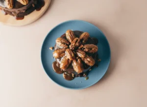 A hero shot of a Cafe Latte Turtle Cake for Two, two small layers of dark fudgy chocolate cake stacked high, drenched in thick glossy dark chocolate and amber caramel sauce dripping down the sides, topped with large glazed toasted pecans, placed on a minimalist white ceramic plate, set on a white marble countertop with a wooden cutting board in the soft background, warm morning light from an east window creating soft shadows and a cozy atmosphere.