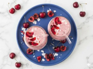 A hero shot of a vibrant pink Cherry Cottage Cheese Smoothie in a clear stemless glass, featuring thick white yogurt swirls visible through the glass. The smoothie is topped with several fresh dark red cherry slices and a sprinkle of snowy white shredded coconut. A clear glass straw is inserted. The scene is set on a white marble countertop with soft morning light from the side, a few whole cherries and coconut flakes are artfully scattered around the base. 4:3 aspect ratio.