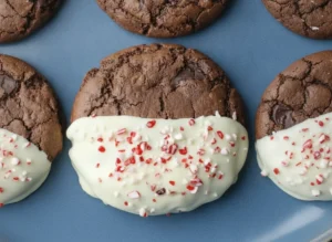 A top-down hero shot of several Chewy Chocolate Peppermint Cookies arranged on a white marble countertop. The cookies are deep dark brown with a cracked, fudgy surface, and each is exactly half-dipped in smooth, creamy white chocolate. The white chocolate portion is generously sprinkled with crushed red and white candy canes. Soft morning light from the east creates gentle shadows. No hands or people. Clean, tidy, and festive presentation.