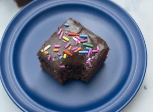 A hero shot of sixteen perfectly cut square chocolate cake bites, each topped with a thick, glossy dark chocolate frosting and a heavy dusting of vibrant rainbow sprinkles. The bites are arranged neatly on a minimalist white ceramic plate sitting on a white marble countertop. Natural morning light from an east-facing window creates soft shadows, with a small wood accent bowl of extra sprinkles and a sprig of fresh mint in the background. 4:3 ratio.