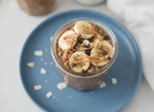 A high-resolution 4:3 hero shot of two elegant glass jars filled with creamy dark chocolate overnight oats. The jars are topped with perfectly sliced circles of fresh banana, a generous scattering of mini dark chocolate chips, and a thick, glossy drizzle of smooth peanut butter flowing down the side. The scene is set on a clean white marble countertop with soft morning sunlight streaming in from an east-facing window, creating gentle shadows. A rustic wooden cutting board and a tiny sprig of fresh mint in a ceramic pot are visible in the soft-focus background. Warm, inviting tones and a minimalist aesthetic.