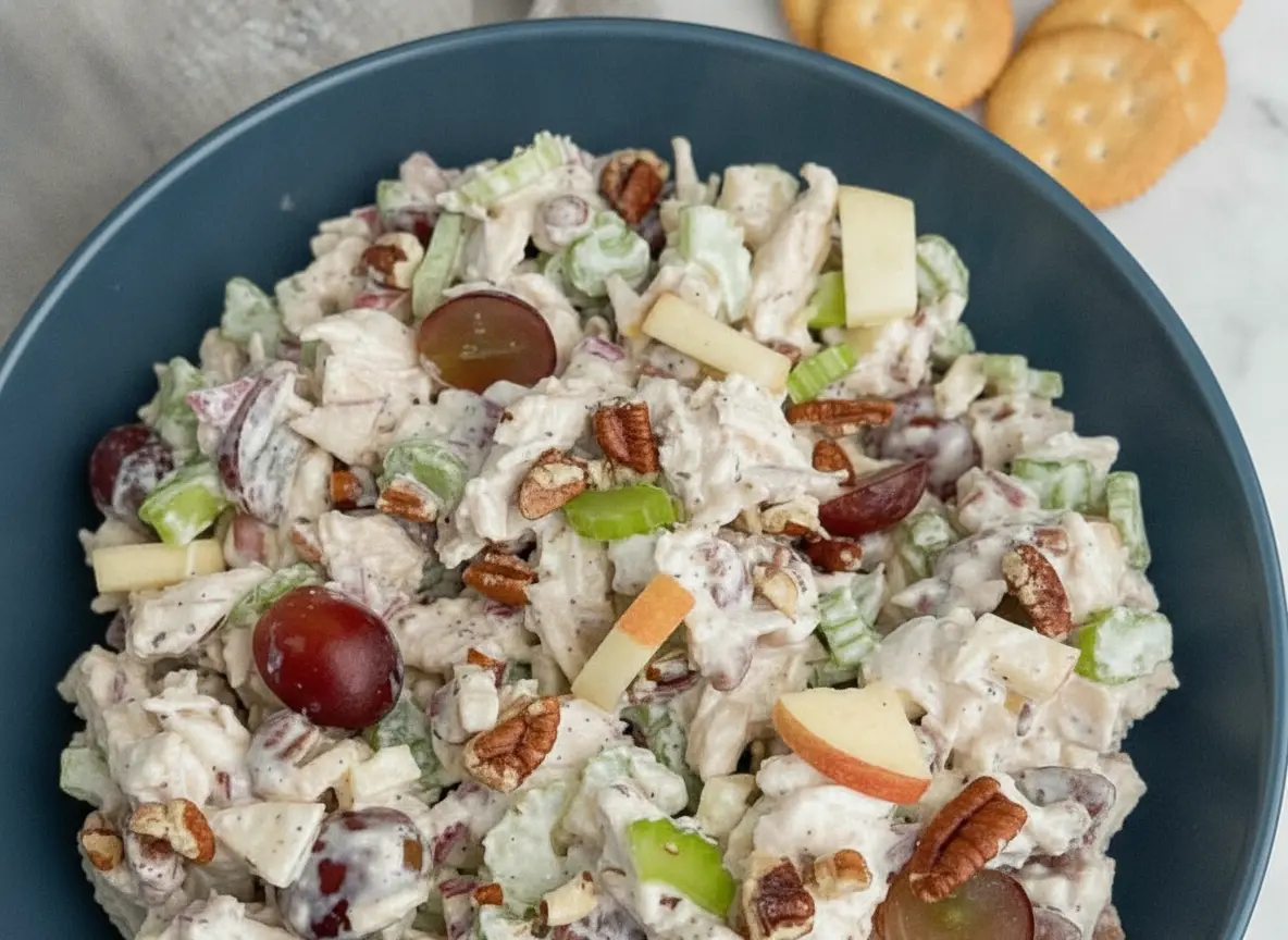 A high-angle 4:3 hero shot of Waldorf Chicken Salad in a minimalist white ceramic bowl placed on a white marble countertop. The salad features shredded white meat chicken, vibrant halved red grapes, thinly sliced green celery, and diced red-skinned apples, all coated in a light creamy dressing and topped with golden chopped pecans. In the background, a few buttery round crackers are scattered on a wooden cutting board. Natural morning light flows in from the side, creating soft, warm shadows.
