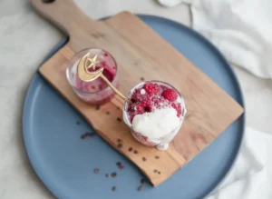 A hero shot of Red Velvet Overnight Oats in a clear glass, showing vibrant magenta and white swirls. The thick white yogurt is dripping artistically over the rim. A gold moon and star spoon is tucked into the oats. Scattered cacao nibs are on the rustic wooden cutting board underneath. Natural morning light coming from a side window onto a white marble countertop.