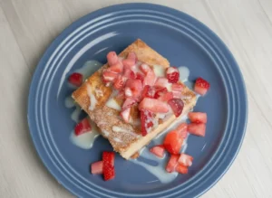 A close-up shot of a single square slice of Crescent Roll Churro Cheesecake on a minimalist white plate. The golden-brown flaky pastry top is dusted with cinnamon sugar, crowned with a mound of fresh, vibrant chopped red strawberries, and drizzled with glossy sweetened condensed milk. Soft morning light from an east window illuminates the scene, casting gentle shadows on a white marble countertop with a rustic wooden cutting board in the background.