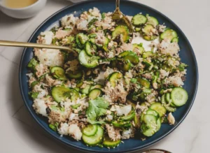 A high-angle hero shot of a Cucumber Crispy Rice Salad Recipe served on a large, minimalist white ceramic platter. The salad features thin, translucent cucumber rounds, jagged golden-brown charred rice clusters, vibrant green cilantro leaves, and scattered crushed peanuts and sesame seeds. Two elegant gold spoons are resting in the salad. The platter sits on a white marble countertop with soft morning light coming from the side, creating gentle shadows. Fresh herbs in a small ceramic bowl are visible in the blurred background. 4:3 aspect ratio.