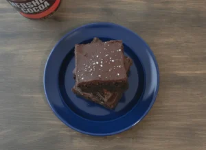 A tall stack of three very dark, almost black, thick fudgy brownies on a minimalist white ceramic plate. The middle brownie shows a thick layer of chocolate frosting between the layers, and the top brownie is crowned with a smooth dark chocolate ganache and generous flakes of sparkling sea salt. The scene is set on a white marble countertop with warm natural morning light from an east window. A dark cocoa tin and a small silver spoon with cocoa powder are blurred in the background. No people, no hands.
