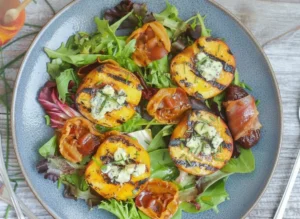 A beautifully composed hero shot of a grilled peach salad in a wide rustic wooden bowl, showcasing charred golden peach halves stuffed with melted crumbled blue cheese, nestled alongside crispy curled pancetta wheels on a bed of mixed baby greens and red radicchio. Finely snipped fresh chives are scattered over the top. The scene is shot on a marble countertop with soft warm morning light coming from an eastern window, casting gentle shadows. A small glass jar of honey with a wooden dipper is visible in the background next to fresh herbs. No hands, no people.
