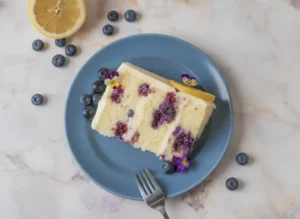 A hero shot of a thick slice of Lemon Blueberry Buttermilk Cake sitting on a minimalist white ceramic plate. The cake has visible layers with white cream frosting and large purple blueberry bursts. A thin lemon wheel and small purple pansies garnish the top. The scene is set on a white marble countertop bathed in soft, natural morning light from an east-facing window, with a wooden cutting board and fresh herbs blurred in the background. 4:3 aspect ratio.