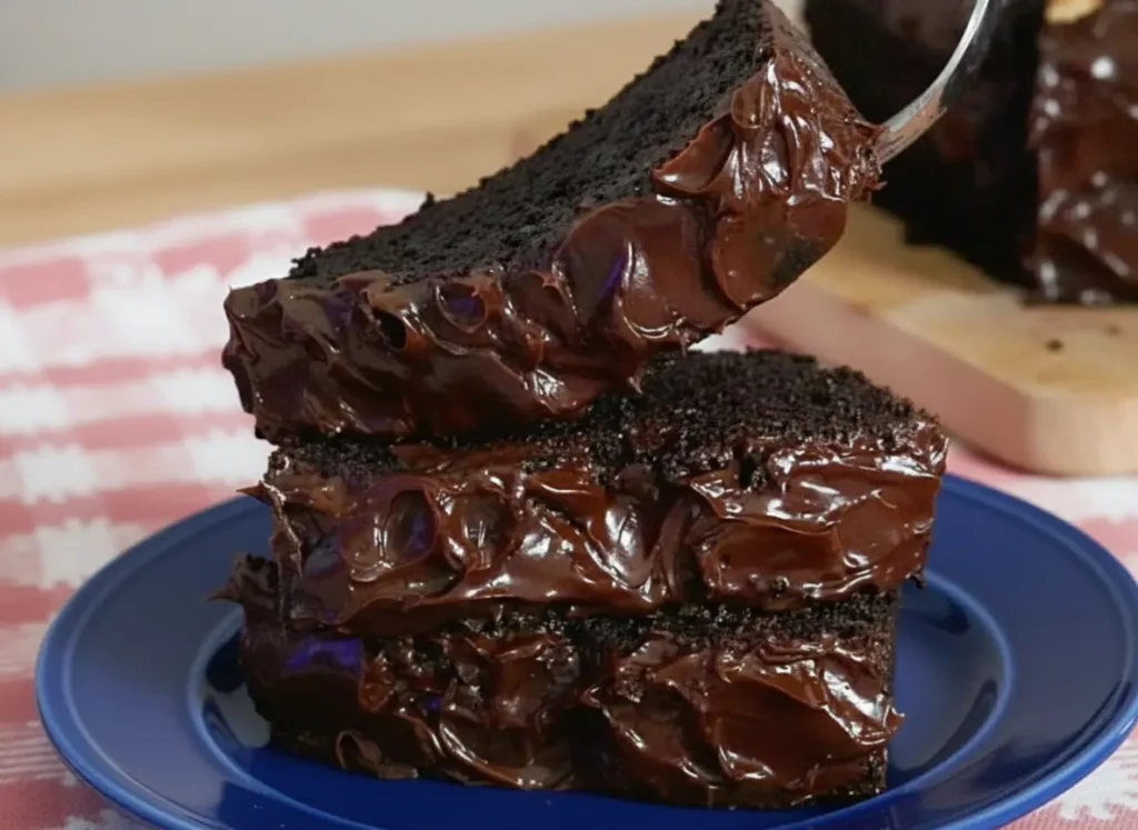 A hero shot of three thick, square slices of Moist Chocolate Banana Cake stacked tall on an orange-peach ceramic plate. Each slice features a dense, dark chocolate crumb and a heavy, thick layer of glossy, swirled chocolate frosting on top. The plate sits on a red-and-white checkered gingham kitchen towel spread over a marble countertop. Bright, warm morning light pours in from an east window, creating soft, natural shadows. In the background, a wooden cutting board and a tiny pot of fresh green herbs are softly out of focus, clean and tidy composition, no hands or people.