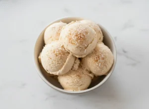 A high-angle hero shot of three scoops of creamy tan No-Churn Coquito Ice Cream with visible cinnamon specks, served in a brown patterned ceramic bowl. The bowl sits on a weathered wooden cutting board on a white marble countertop. In the background, a whole coconut and green tropical leaves are visible. Soft morning light enters from the side, creating warm tones and gentle shadows. 4:3 aspect ratio.