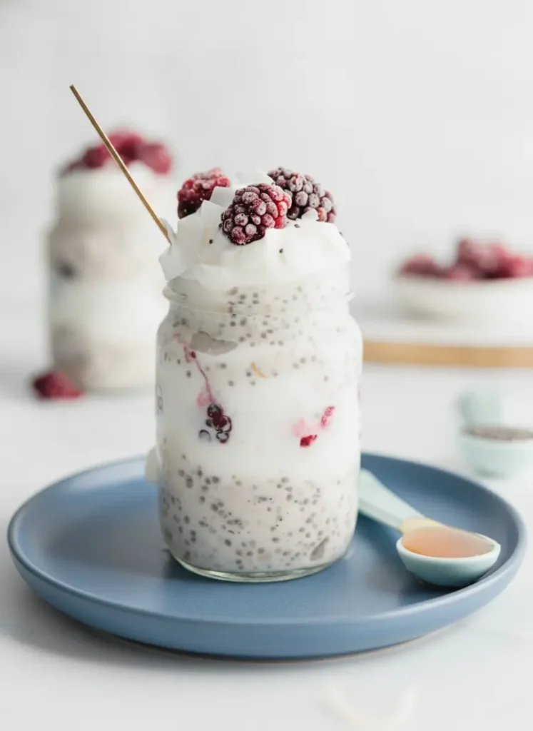 Protein Overnight Oats With Coconut Milk A top-down prep shot on a weathered wooden cutting board. A minimalist white ceramic bowl contains dry rolled oats, a small pile of black chia seeds, and a scoop of fine vanilla protein powder. A splash of thick white coconut milk is being poured from a small pitcher, showing the rich texture. The background features a clean marble surface and a hint of fresh green mint leaves.