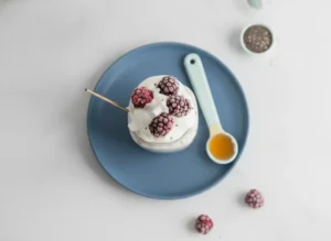 A hero shot of a mason jar filled with creamy white protein overnight oats, showing visible chia seeds and swirls of purple juice from frozen blackberries. The jar is topped with frosty blackberries, raspberries, and large white coconut flakes. A thin wooden stir stick rests inside the jar. The setting is a bright marble countertop in an east-facing kitchen with soft morning sunlight casting gentle shadows. A small ceramic dish of honey sits nearby on the white marble.