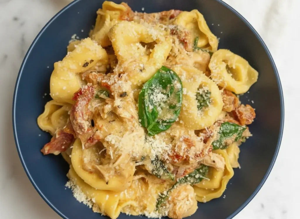 A hero shot of Slow Cooker Marry Me Tortellini in a minimalist white ceramic bowl. The pasta is coated in a rich, creamy orange-tinted sauce with visible strips of red sun-dried tomatoes and bright green spinach leaves. A gold spoon is resting in the bowl, lifting a single tortellini. The bowl sits on a white marble countertop with a soft morning light coming from the side, creating gentle shadows. Fresh basil leaves and a wooden cutting board are blurred in the background. 4:3 aspect ratio.