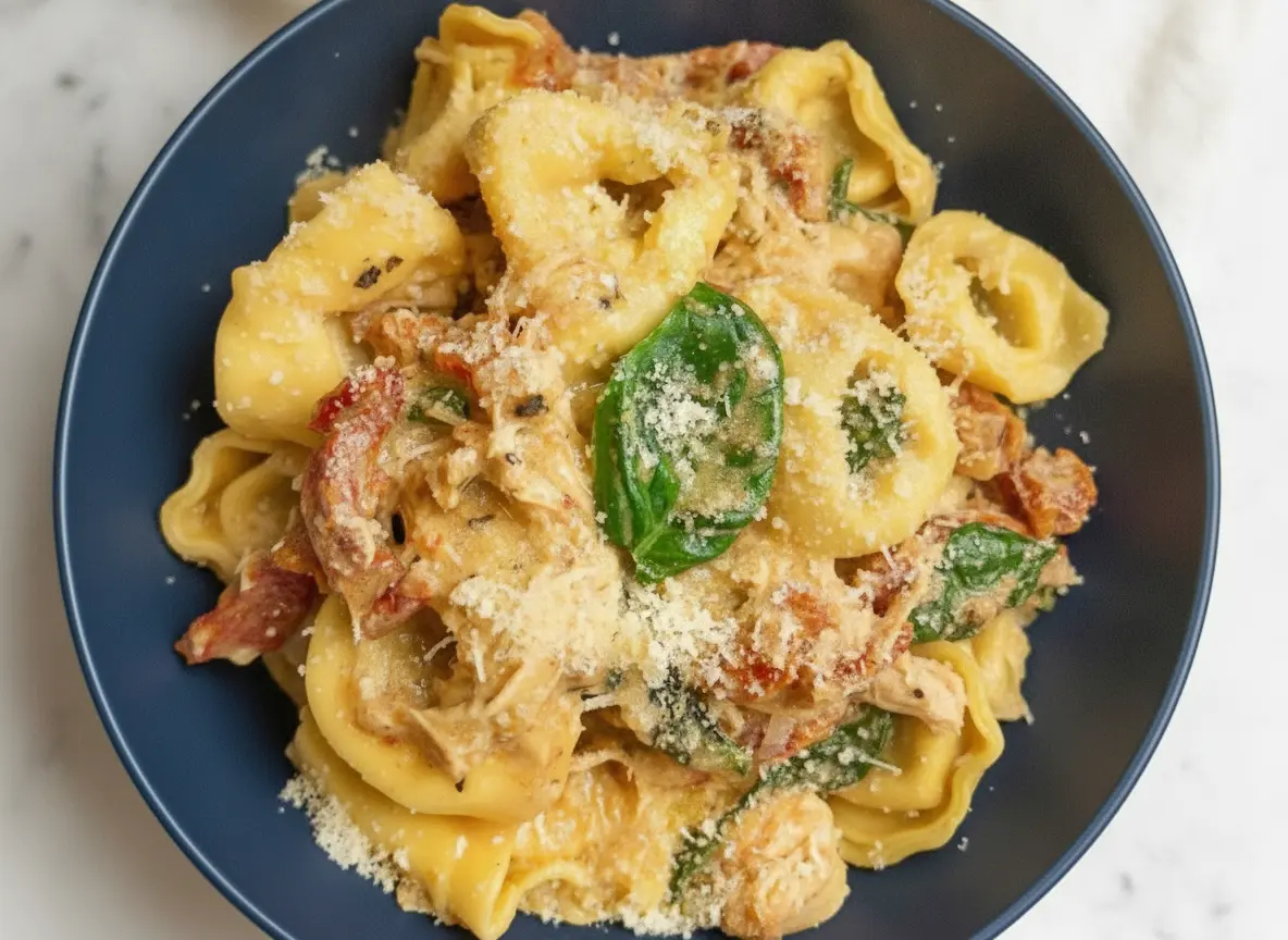 A hero shot of Slow Cooker Marry Me Tortellini in a minimalist white ceramic bowl. The pasta is coated in a rich, creamy orange-tinted sauce with visible strips of red sun-dried tomatoes and bright green spinach leaves. A gold spoon is resting in the bowl, lifting a single tortellini. The bowl sits on a white marble countertop with a soft morning light coming from the side, creating gentle shadows. Fresh basil leaves and a wooden cutting board are blurred in the background. 4:3 aspect ratio.