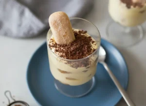A hero shot of two elegant glasses filled with Tiramisu For Two, placed on a marble countertop with soft morning light from an east window. The tiramisu has visible layers of pale yellow mascarpone cream and coffee-soaked ladyfingers. The top is dusted with dark cocoa powder and one whole ladyfinger is tucked vertically into the cream. One glass sits on a terracotta-colored ceramic plate with a small silver spoon. A grey linen napkin is blurred in the background next to a wooden cutting board. Warm tones and soft shadows.