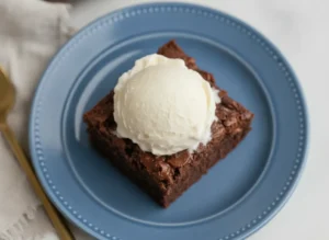 A hero shot of a single thick slice of Chocolate Crunch Cake on a minimalist white ceramic plate. The cake has a dark, moist crumb and a heavy, jagged topping of chocolate-coated cereal shards. A large, perfectly round scoop of white vanilla ice cream is melting over the top, its creaminess dripping into the chocolate crevices. The plate sits on a white marble countertop with a weathered wooden cutting board partially visible. Soft, warm morning light from an east window creates gentle shadows. 4:3 aspect ratio.