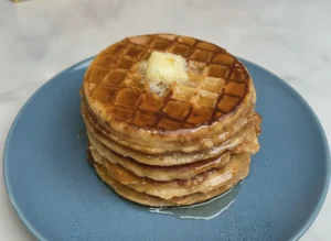 A high-angle 4:3 hero shot of a stack of four thick, golden-brown Viral French Toast Waffles. The waffles have visible cinnamon flecks and deep pockets. A large, melting pat of yellow butter sits on top, with thick amber maple syrup dripping down the sides and pooling on a minimalist white ceramic plate. The scene is set on a white marble countertop with soft morning light from a window creating gentle shadows. Fresh mint is visible in the soft-focus background.