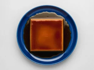 A professional hero shot of a rectangular Dulce de Leche Crema Volteada (Flan) on a minimalist white ceramic plate with a textured rim. The flan is tall and perfectly shaped, topped with a glossy, dark mahogany caramel glaze that pools into a deep amber sauce around the base. The scene is set on a white marble countertop with soft morning light from an east window creating gentle shadows. A small wooden cutting board and a sprig of mint are visible in the soft-focus background.