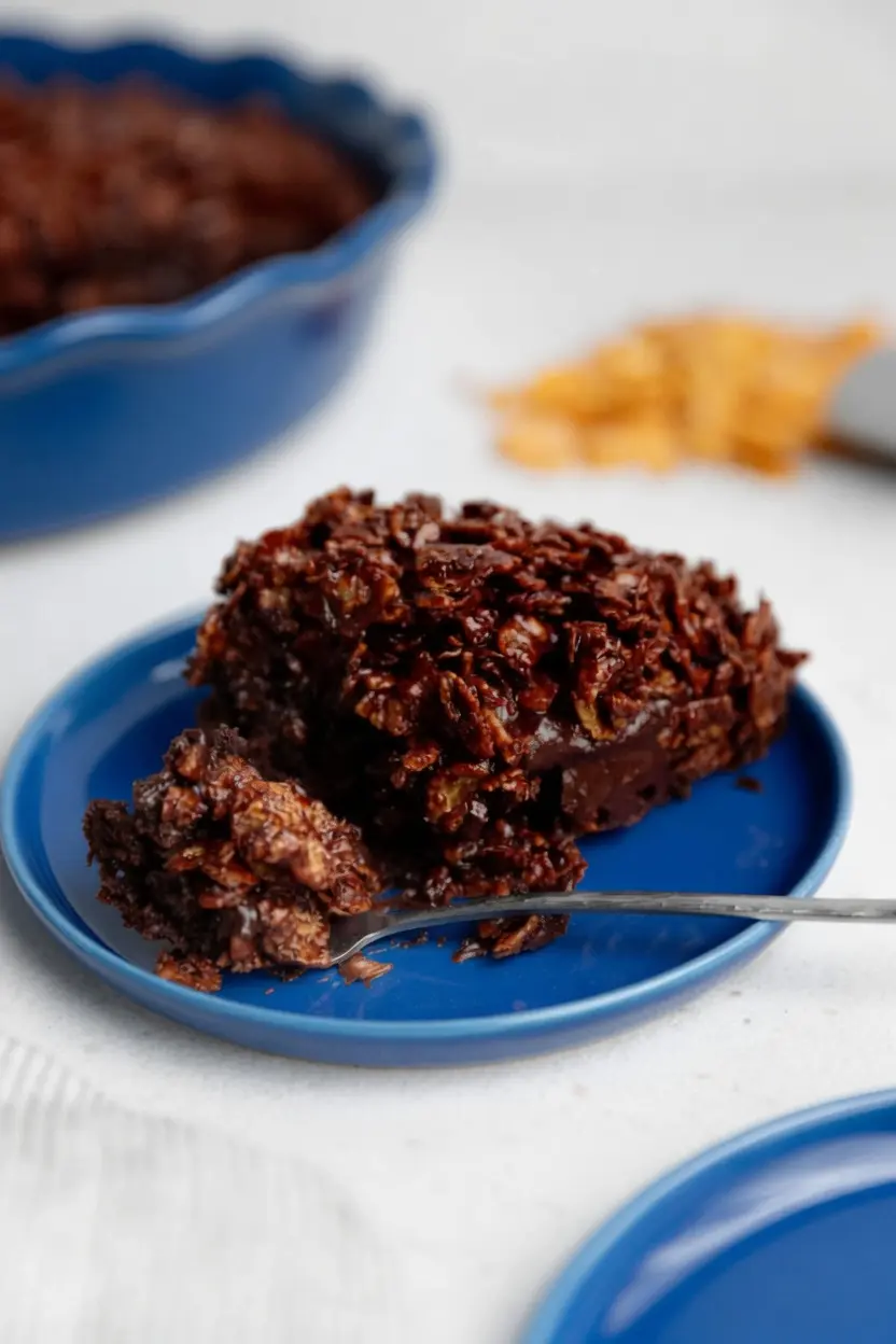 Close-up of the technique showing the chocolate-coated cereal being spread over the fudgy cake base inside a white ceramic baking dish. The texture of the glistening chocolate glaze on the flakes is highly detailed. No hands visible. 3:4 aspect ratio.