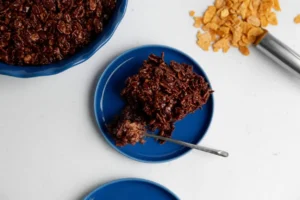 A hero shot of a Chocolate Crunch Cake on a minimalist white plate, sitting on a white marble countertop with warm morning light from an east window. The cake has a deep, dark fudgy base and a thick, irregular topping of dark chocolate-coated cornflakes. A silver fork has just taken a bite, revealing the soft interior. In the background, a wooden cutting board and a small pot of fresh mint are softly blurred. 4:3 aspect ratio, soft shadows, warm tones.